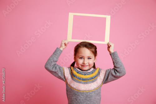 Portrait of a little smiling girl in warm dress holding blank frame for mock-up on a pink background