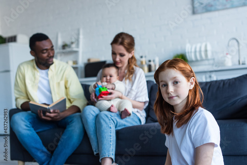 redhead preteen girl looking at camera near multiethnic family sitting on sofa on blurred background