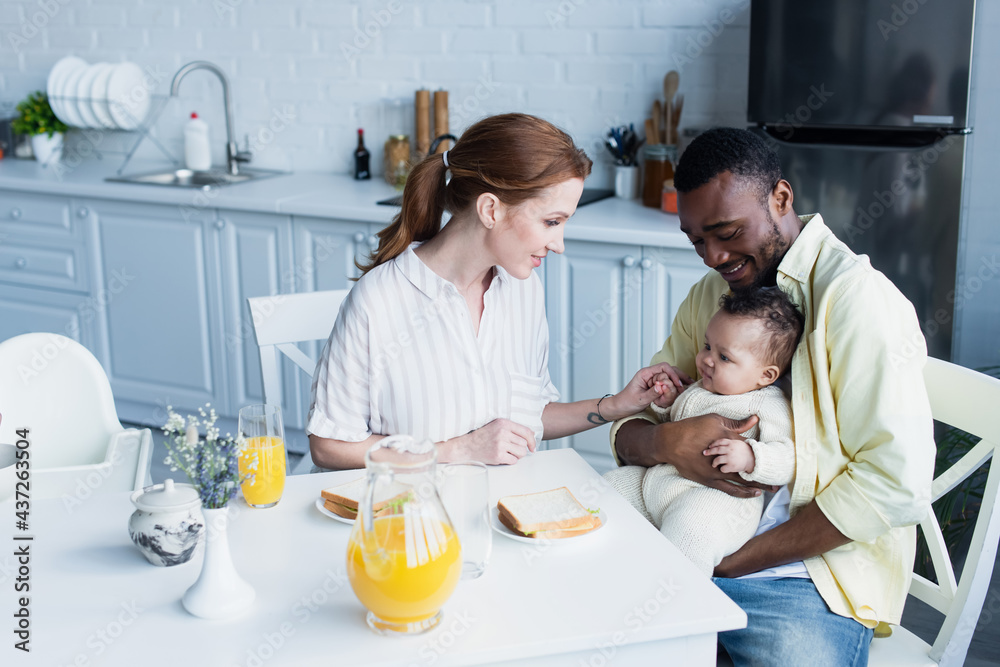 happy african american man holding baby near smiling wife during breakfast happy african american man holding baby near smiling wife during breakfast