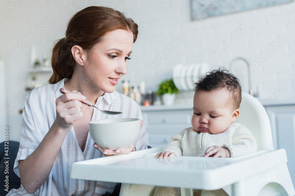 smiling woman holding bowl and spoon near infant african american girl sitting in baby chair smiling woman holding bowl and spoon near infant african american girl sitting in baby chair