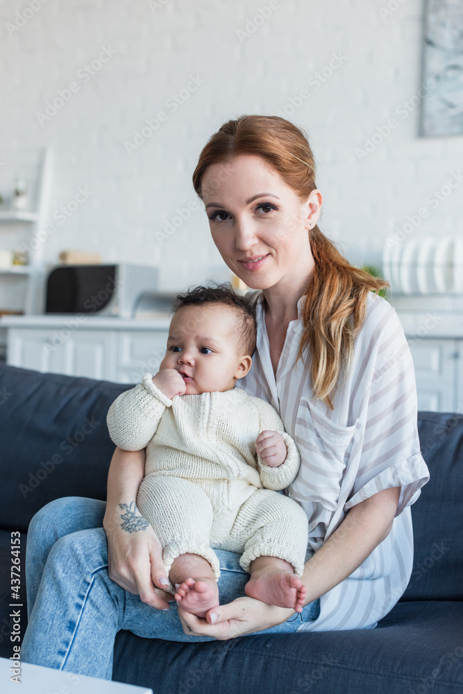 smiling woman looking at camera while sitting on sofa with little african american daughter smiling woman looking at camera while sitting on sofa with little african american daughter