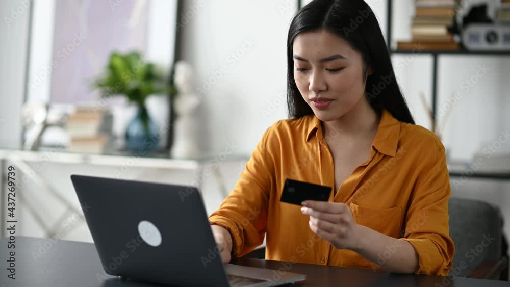 Online shopping. Happy beautiful young asian brunette woman sitting at the table, uses laptop and credit card to pay for online purchases and delivery, enters data, looks at the card, smiles Online shopping. Happy beautiful young asian brunette woman sitting at the table, uses laptop and credit card to pay for online purchases and delivery, enters data, looks at the card, smiles
