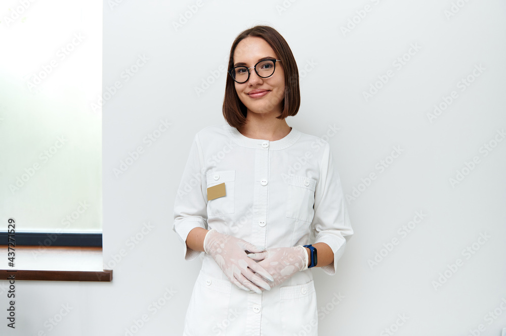 Confident portrait of a young doctor in white medical lab coat and protective gloves looking at camera while posing against white wall in cosmetology clinic