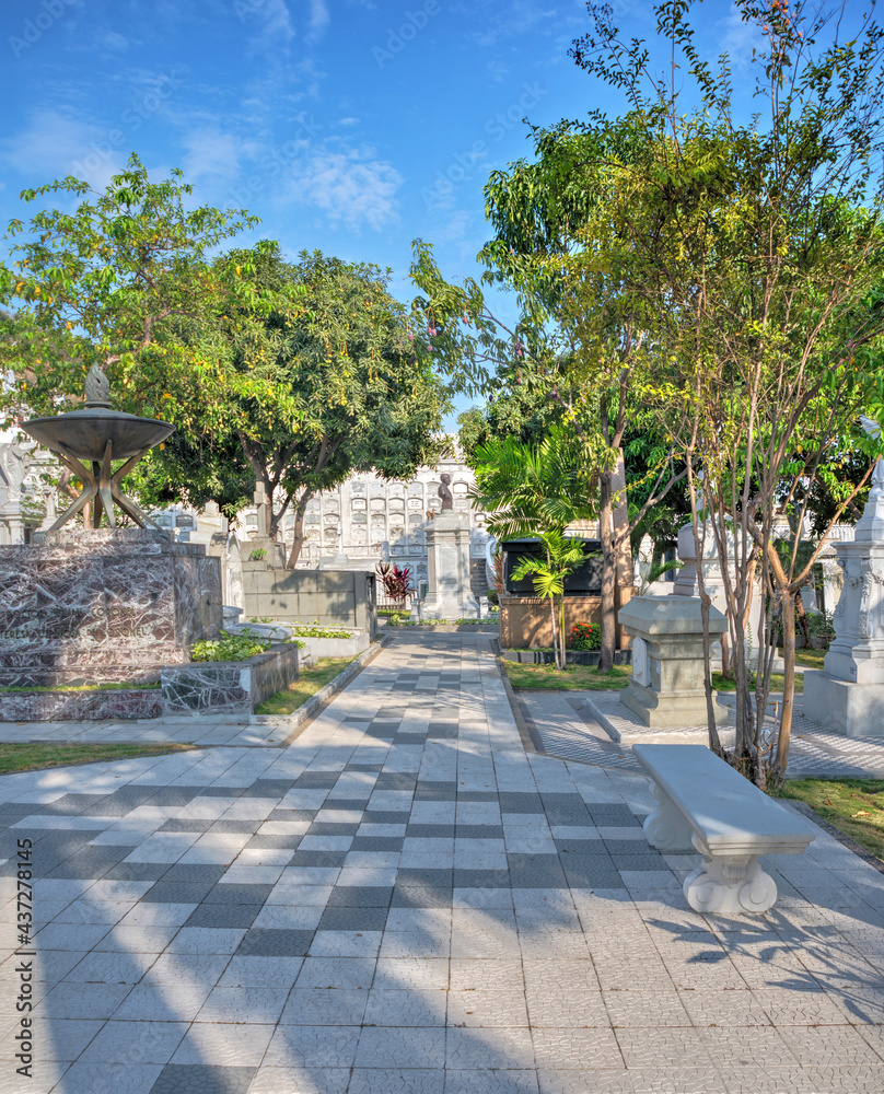 Section of a cemetery in Guayaquil, Ecuador, with a nice tile floor design, trees, benches and statues, on a sunny summer afternoon