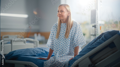 Modern Hospital Ward: Beautiful Caucasian Female Patient Resting on a Bed, Fully Recovering after Successful Surgery, Sickness or Coronavirus she Smiles. Behind Her Window with a Beautiful Sunny View