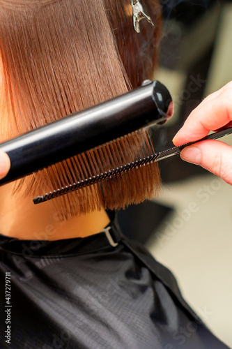 Hair stylist s hands straightening short hair of young brunette woman with flat iron and comb in a beauty salon