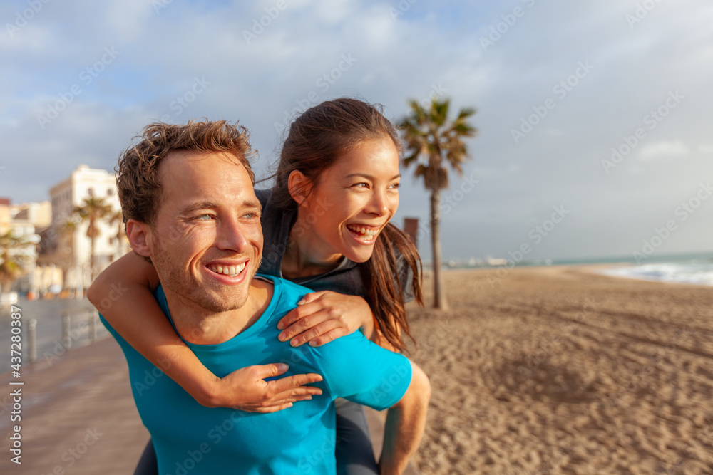 Happy interracial couple having fun laughing during summer vacation. Portrait of young Asian woman piggybacking on male Caucasian boyfriend on Europe travel beach, Barcelona. Happy interracial couple having fun laughing during summer vacation. Portrait of young Asian woman piggybacking on male Caucasian boyfriend on Europe travel beach, Barcelona.