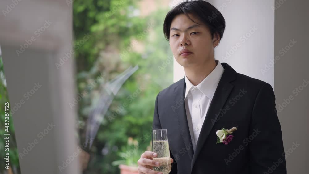 Young groom looking out the window turning to camera. Portrait of sad Asian man in wedding suit holding champagne glass thinking. Unhappy guy forced getting married