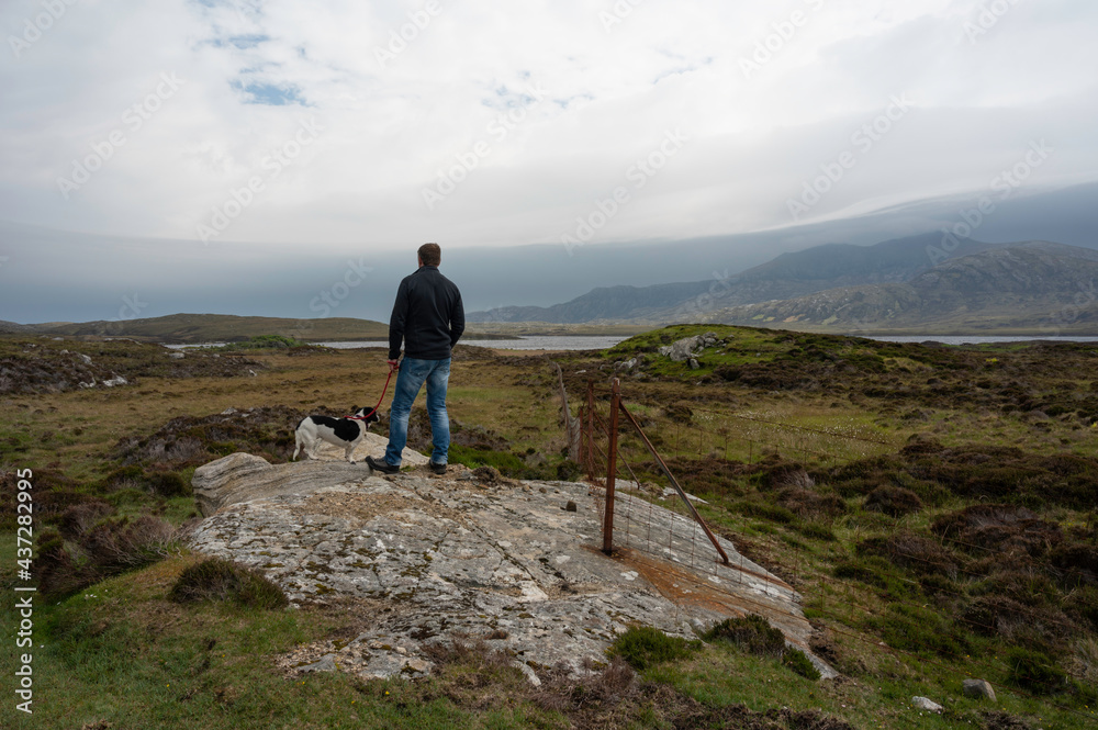 Caucasian man with small black and white dog looking out over Loch Druidibeag nature reserve in South Uist. Taken from behind. Rock, fence, loch, grass, mountains in view. Dramatic clouds. Caucasian man with small black and white dog looking out over Loch Druidibeag nature reserve in South Uist. Taken from behind. Rock, fence, loch, grass, mountains in view. Dramatic clouds.