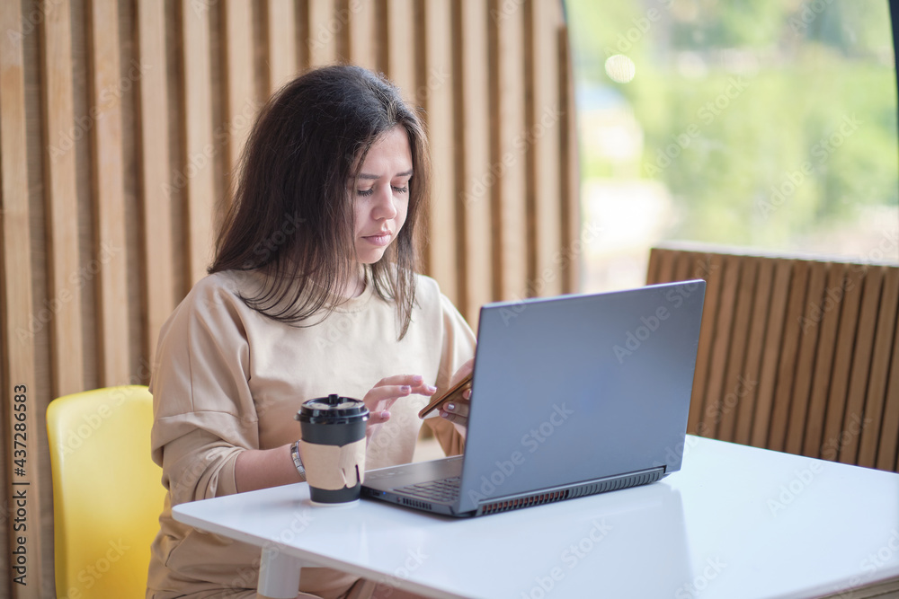 Female holding phone and looking on his screen. Young hipster woman sitting in coffee, drinking coffee and using laptop.On table is laptop. Girl browsing internet, chatting, blogging. Female holding phone and looking on his screen. Young hipster woman sitting in coffee, drinking coffee and using laptop.On table is laptop. Girl browsing internet, chatting, blogging.