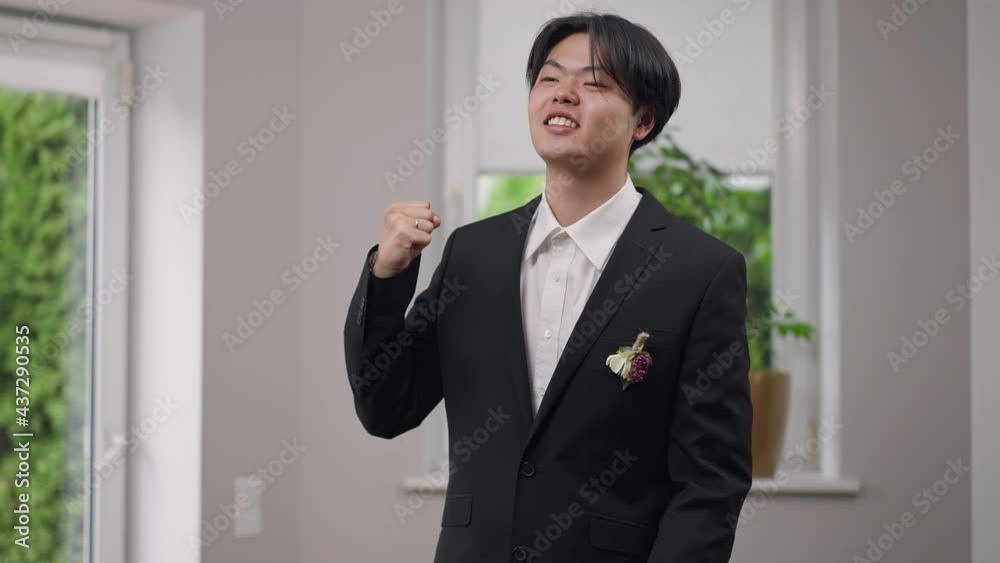 Cheerful Asian groom with toothy smile showing victory gesture looking away laughing. Portrait of happy excited young man in elegant suit rejoicing on wedding morning indoors. Marriage and happiness Cheerful Asian groom with toothy smile showing victory gesture looking away laughing. Portrait of happy excited young man in elegant suit rejoicing on wedding morning indoors. Marriage and happiness