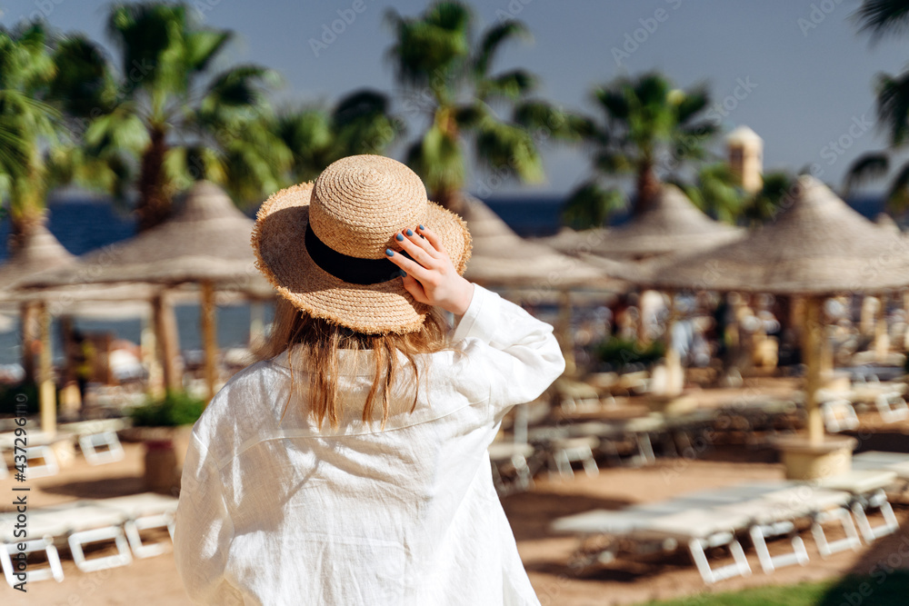 View from the back, a girl in a straw hat on the beach near the sea, among beach chairs and umbrellas. Young girl resting in a warm country