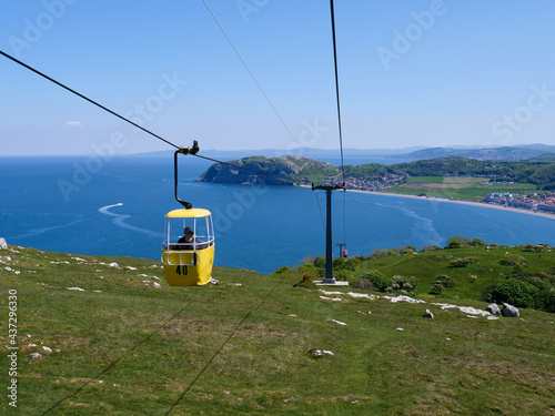 Yellow color overhead cable car with two unrecognizable people in it moving up from tourist resort town Llandudno to Great Orme hill top.