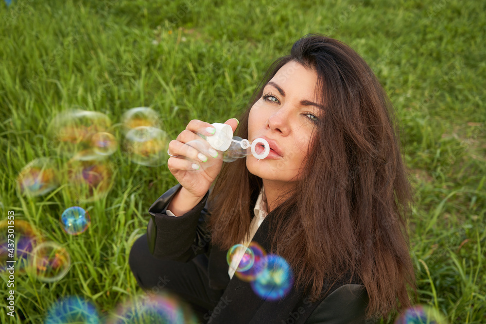A young beautiful woman with dark long hair is sitting on the green grass blowing soap bubbles. A young beautiful woman with dark long hair is sitting on the green grass blowing soap bubbles.