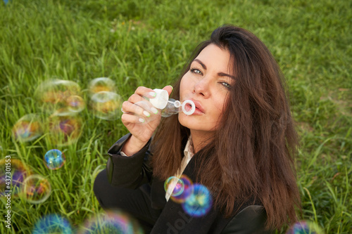 A young beautiful woman with dark long hair is sitting on the green grass blowing soap bubbles.