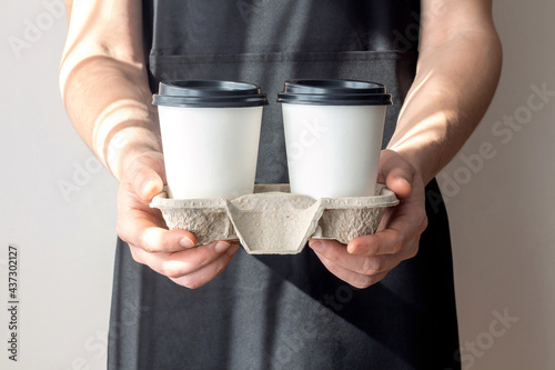 A man holding disposable paper coffee cups in a cardboard tray. A person in a black apron with takeout mugs.