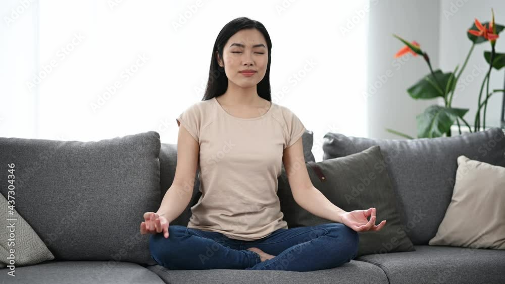 Calm beautiful young asian brunette woman, in casual clothes, sitting at home on the sofa in the lotus position, resting, taking care of her health, relaxing and meditating, closing her eyes Calm beautiful young asian brunette woman, in casual clothes, sitting at home on the sofa in the lotus position, resting, taking care of her health, relaxing and meditating, closing her eyes