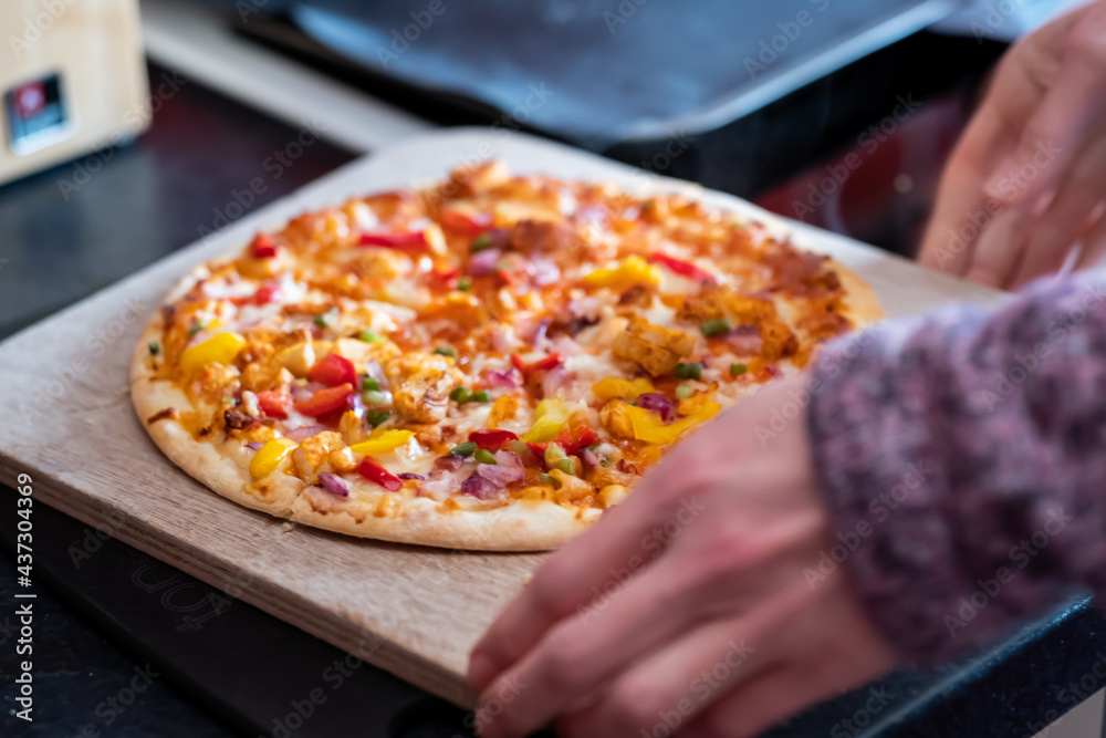 Close up and selective focus of a home cooked thin crust sweet chilli chicken pizza on a wooden cutting board being sliced with a pizza roller by an unidentifiable woman