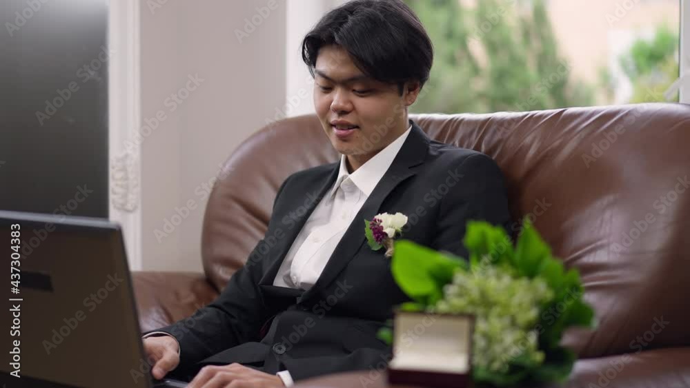 Medium shot of smiling Asian groom in wedding suit with boutonniere typing on laptop keyboard sitting on armchair indoors. Happy young man in costume messaging online on wedding morning Medium shot of smiling Asian groom in wedding suit with boutonniere typing on laptop keyboard sitting on armchair indoors. Happy young man in costume messaging online on wedding morning