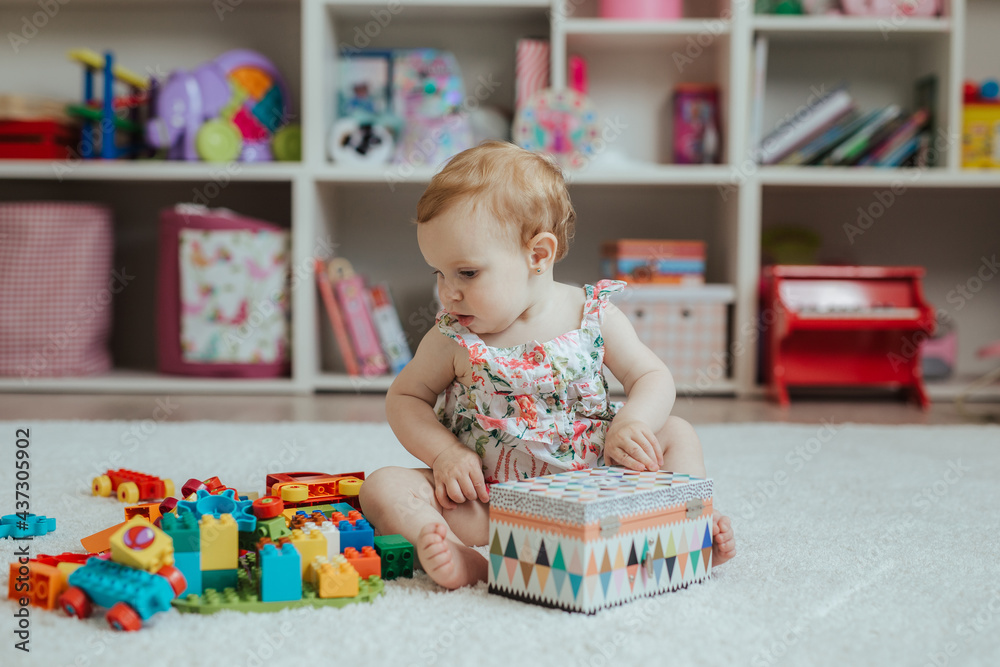 Little curious baby girl playing on a playground in a bright sunny room. Learning and playing colorful educational blocks game and puzzle Little curious baby girl playing on a playground in a bright sunny room. Learning and playing colorful educational blocks game and puzzle