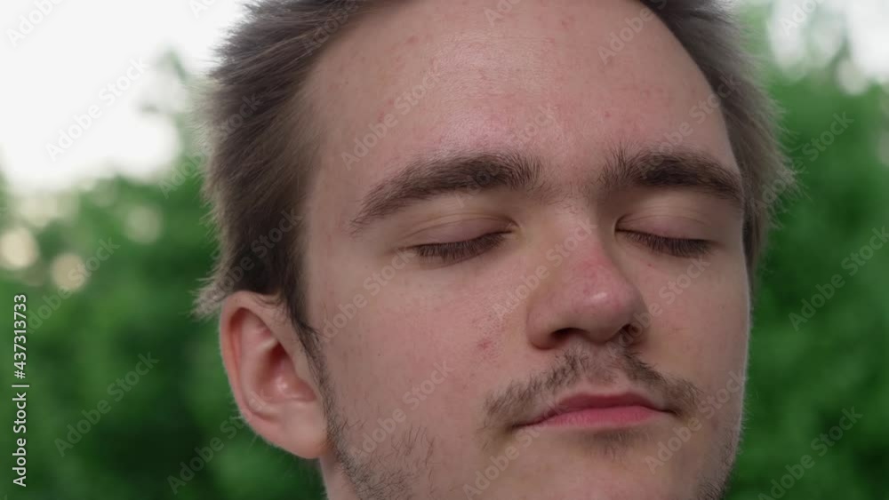 The face of a young handsome guy is shot very close, he stands with his eyes closed and thinks about something, he opens his eyes and looks at the camera behind against the background of green foliage The face of a young handsome guy is shot very close, he stands with his eyes closed and thinks about something, he opens his eyes and looks at the camera behind against the background of green foliage
