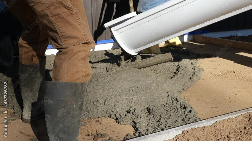 Cement concrete is poured within a wood form to make a new sidewalk next to a building. Working men in rubber boots use rakes to spread the wet mix to keep the thickness even throughout. Two clips. Cement concrete is poured within a wood form to make a new sidewalk next to a building. Working men in rubber boots use rakes to spread the wet mix to keep the thickness even throughout. Two clips.