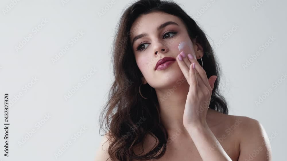 Skin moisturizing. Facial care. Beauty wellness. Brunette woman with bare shoulders applying cream on radiant face isolated on light neutral copy space background. Skin moisturizing. Facial care. Beauty wellness. Brunette woman with bare shoulders applying cream on radiant face isolated on light neutral copy space background.