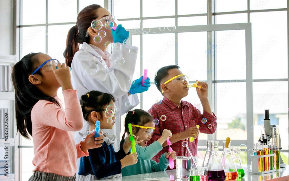 Asian female scientist teacher in white lab coat having fun playing blowing soap bubbles with little innocent happy pre-elementary boy and girl children wears safety goggles in school laboratory - obrazy, fototapety, plakaty 