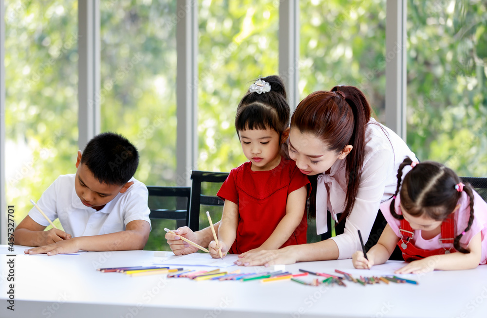 Fototapeta premium Asian female pretty single mother standing helping teaching little cute innocent daughter drawing and painting with colored pencils on paper book while other boy and girl doing homework together