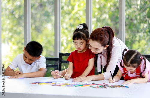 Asian female pretty single mother standing helping teaching little cute innocent daughter drawing and painting with colored pencils on paper book while other boy and girl doing homework together