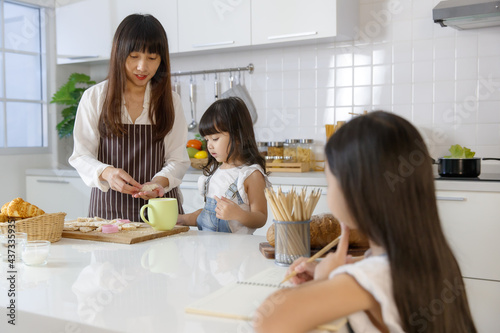 A cute little 7 years old Asian girl sitting and doing homework in kitchen while her mother teaching her younger sister how to make food and cooking behind.