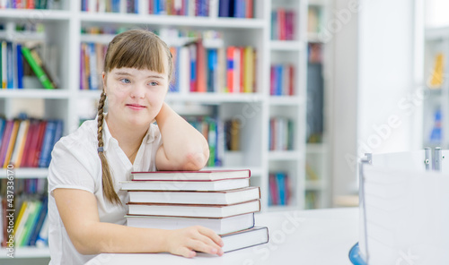 Pensive young girl with syndrome down sita at library with books. Empty space for text. Education for disabled children concept