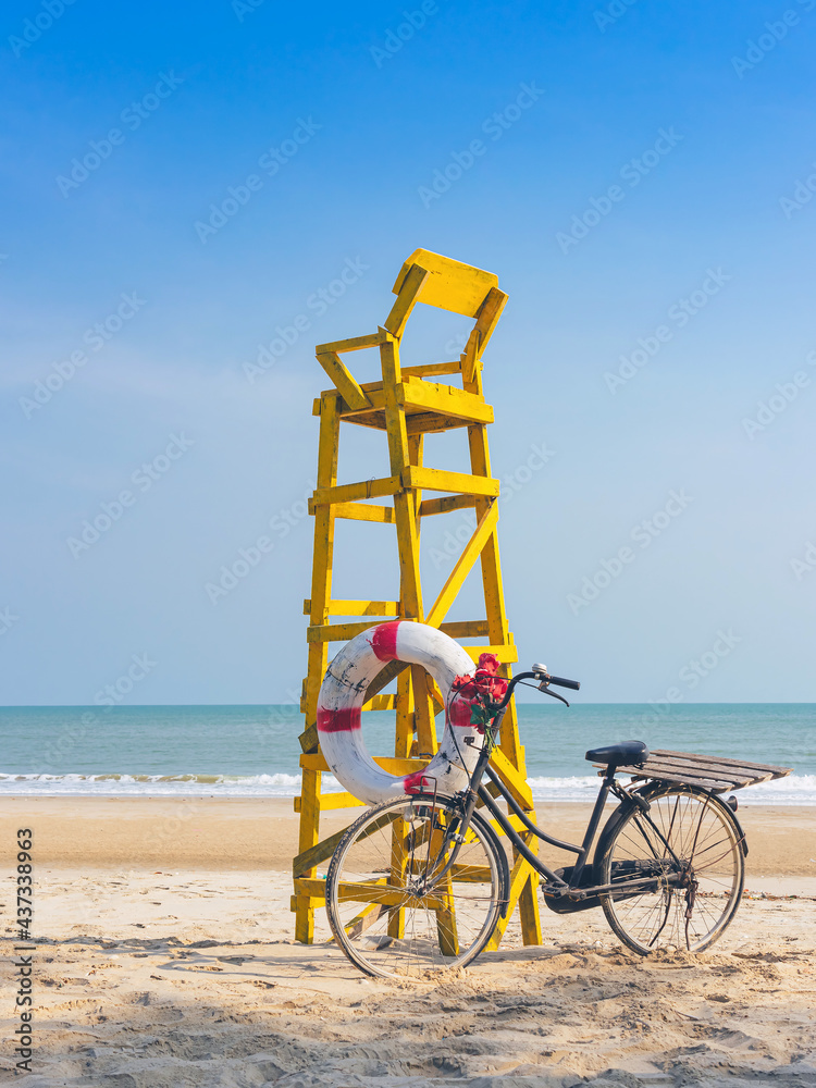 Fototapeta premium Old retro black bicycle with flowers bouquet beside Life ring for life safety on yellow lifeguard stand station or lifeguard chair protecting the safety of tourist on the empty beach in sunshine.