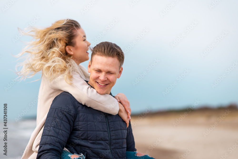 Happy man giving piggyback ride to his woman and laughing at beach. Smiling guy in love carrying on back her girlfriend.