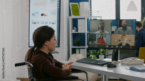 Paralyzed immobilized manager woman speaking online with remotely coworkers sitting wheelchair in start up busineess office at computer. Handicapped employee talking on virtual meeting photo