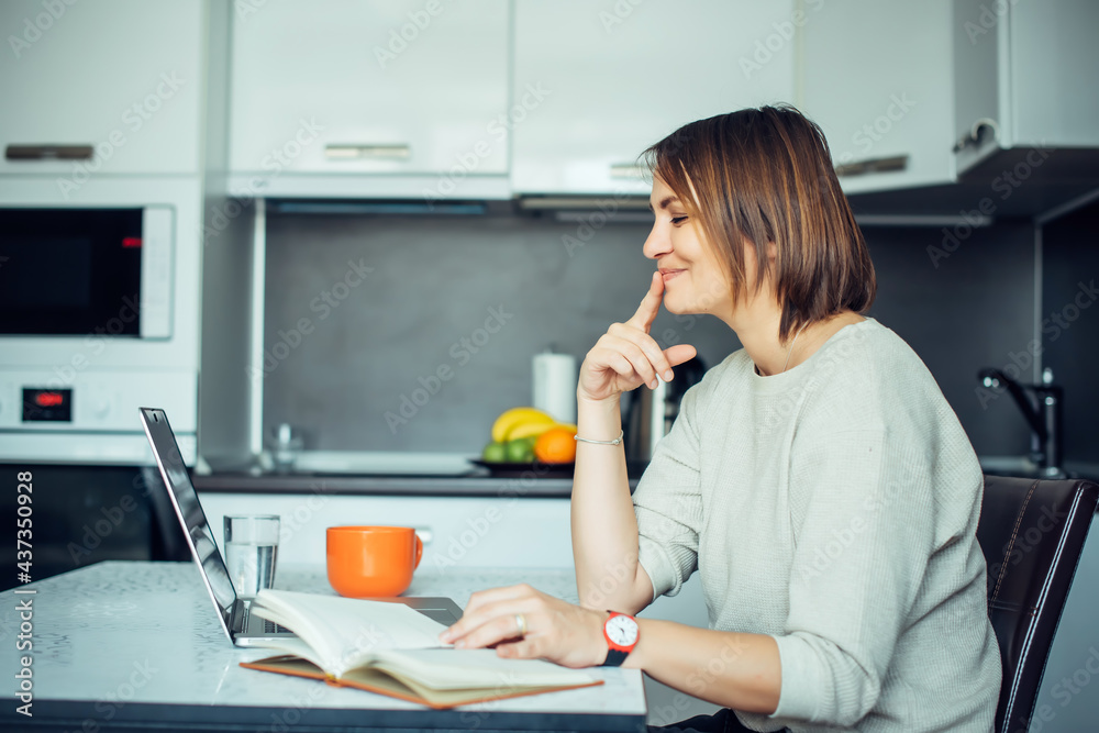 Young pretty brunette with laptop sitting at a table in the kitchen. Female writer works at home and drinks coffee. Freelance, remote work, creativity. Young pretty brunette with laptop sitting at a table in the kitchen. Female writer works at home and drinks coffee. Freelance, remote work, creativity.