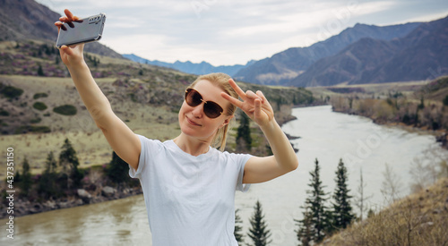 Young blonde girl in sunglasses and white T-shirt takes a selfie on her smartphone on the background of river and rocky mountains. Happy female tourist smiles at mobile phone camera.