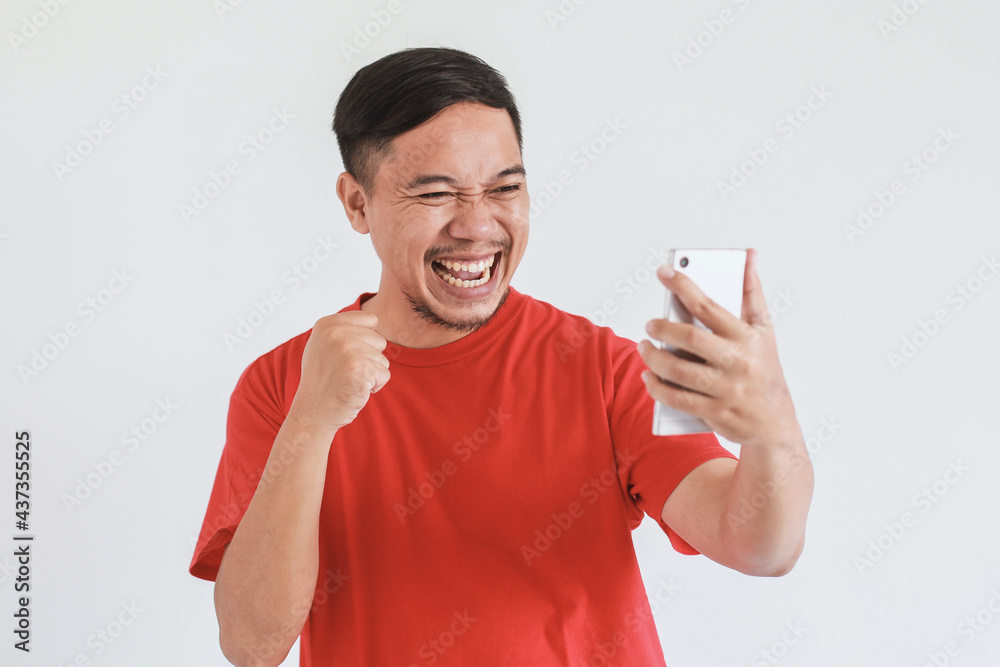 Smiling face expression of happy Asian man wearing red t-shirt looking to the smart phone with fist hand Smiling face expression of happy Asian man wearing red t-shirt looking to the smart phone with fist hand