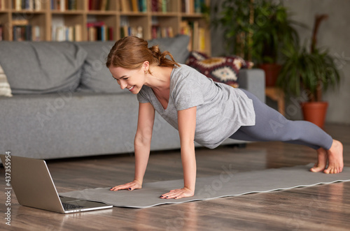 Happy sporty woman standing in plank pose in front of laptop, doing yoga online