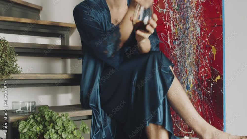 Handheld camera shot of barefoot transgender man in silk robe sitting on wooden stairs at home, drinking alcohol cocktail and smoking cigarette