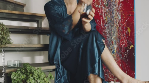 Handheld camera shot of barefoot transgender man in silk robe sitting on wooden stairs at home, drinking alcohol cocktail and smoking cigarette