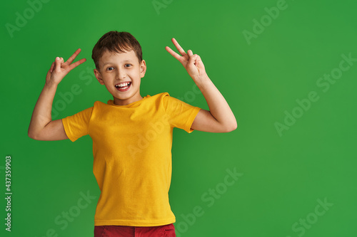 Portrait happy and victorious boy in yellow T-shirt showing a gesture of victory and peace, smiling, making a v sign, with both hands, a cute child enjoying victory in the studio on a green background
