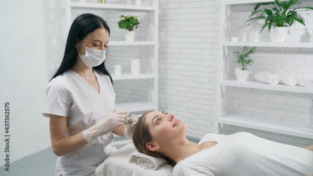 Side view of brunette beautician in protective mask doing injection for young attractive woman in hair. Concept of improvements hair with special cosmetics. Side view of brunette beautician in protective mask doing injection for young attractive woman in hair. Concept of improvements hair with special cosmetics.