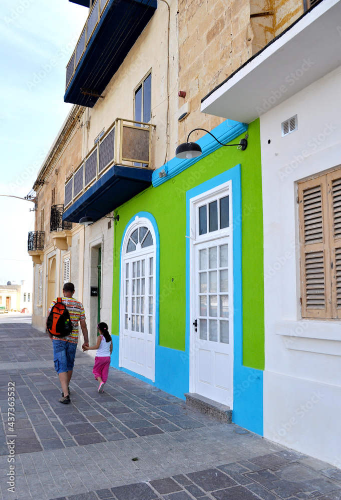 A father and his daughter are walking down a street in Marsaxlokk with a beautiful old house with a bright green and blue painted wall. Malta, Europa