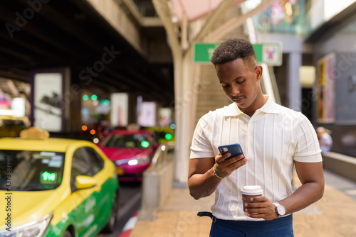 Portrait of young African businessman wearing casual clothes while using mobile phone and holding coffee cup at taxi station in city