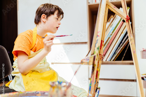 Education and special child concept. Artist school boy in apron painting brush watercolors portrait on a easel.