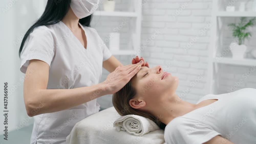 Side view of young brunette beautician doing face massage for young beautiful woman in beauty salon. Concept of special procedures for improvement skin face and all body. Side view of young brunette beautician doing face massage for young beautiful woman in beauty salon. Concept of special procedures for improvement skin face and all body.
