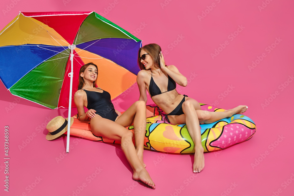 Cheerful girls in swimsuits on inflatable mattress under sun umbrella on pink studio background