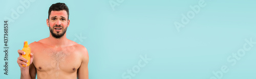 Worried shirtless man with sunburn holding sunscreen isolated on blue, banner photo
