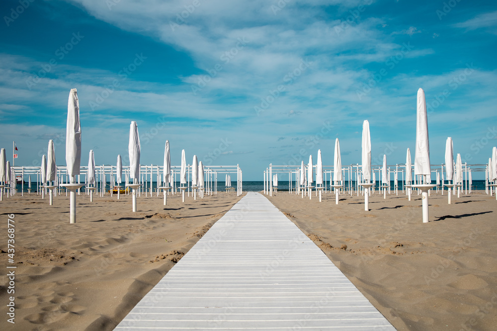 Riviera Romagnola spring view. Almost summer, clue skies, white walking path, sunbeds and umbrellas ready for season opening. Italian vintage riviera beach on the adriatic coast.  - obrazy, fototapety, plakaty 